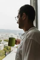 Person looking peacefully out window at sunrise holding warm coffee cup
