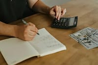 Handwritten budget notes on paper next to calculator and coffee mug